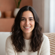 Portrait of a smiling Spanish woman in her late 20s with dark hair, wearing casual clothing, against a warm, blurred backgrou